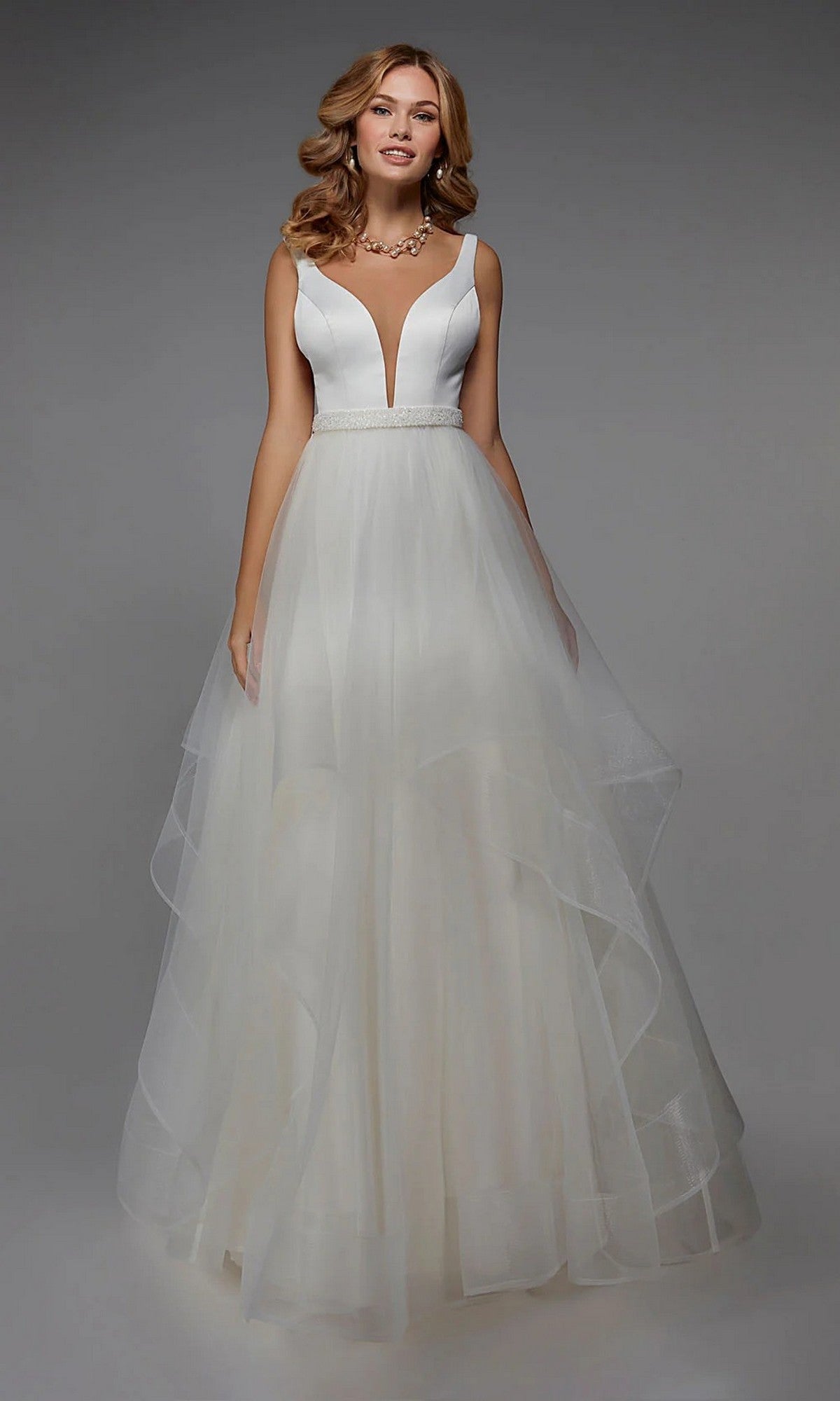 A woman wears the Alyce 7022 long ivory dress, featuring a sleeveless deep V neckline, fitted bodice, beaded waistband, and layered tulle skirt, standing against a plain gray background.
